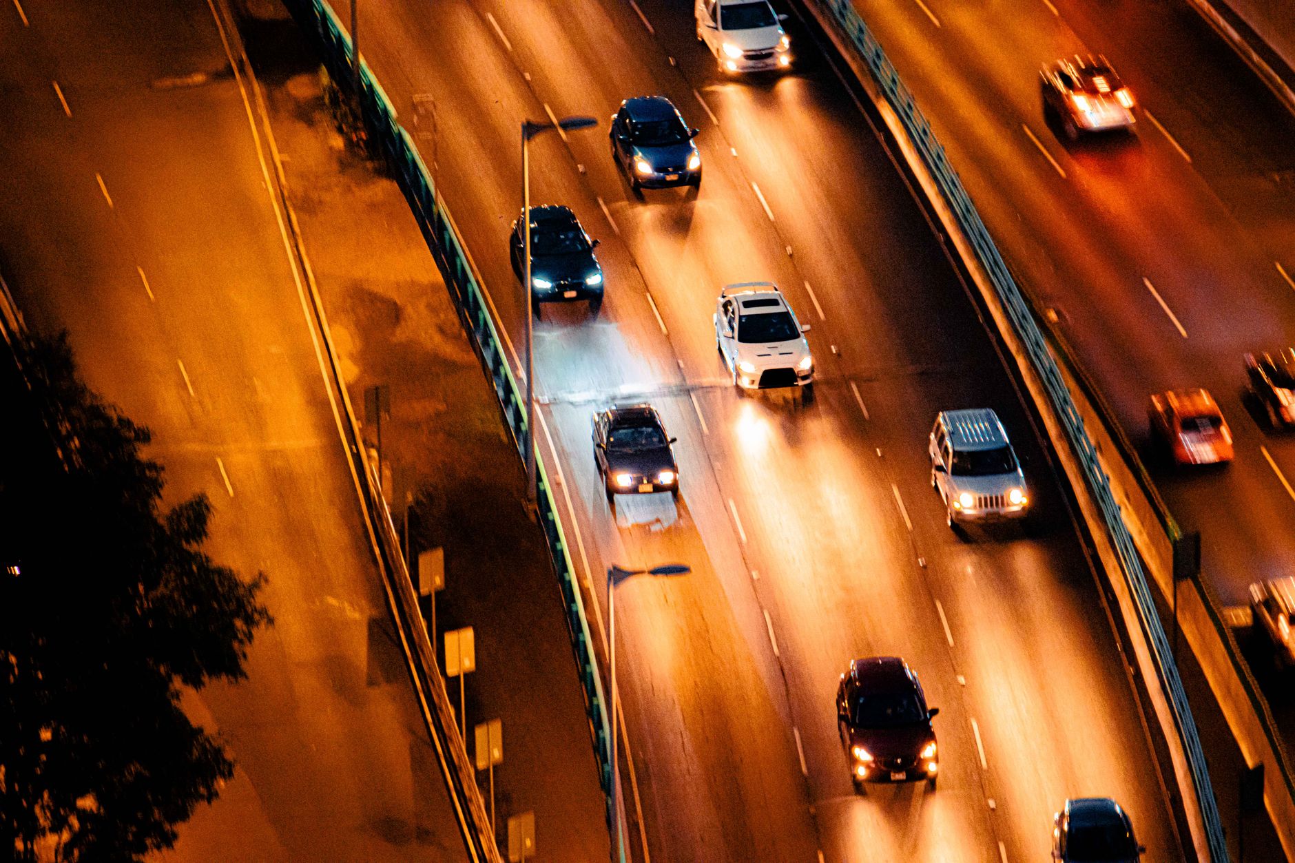 Aerial night view of cars with headlights on driving along a multi-lane highway lit by orange street lights.