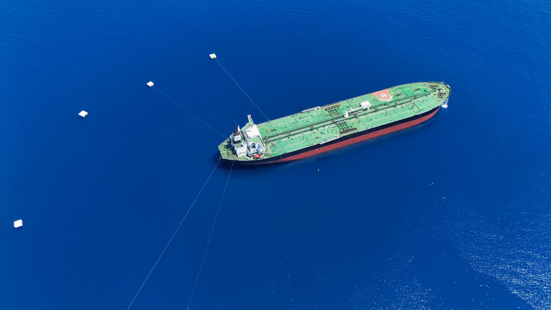 Aerial view of a large oil tanker with a green deck and red hull, held by mooring lines to white buoys on deep blue sea.